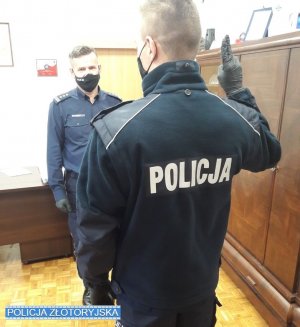 Nowy policjant stoi twarzą zwrócony do Komendanta. Prawą rękę ugiętą w łokciu trzyma uniesioną do góry jak przy śłubowaniu.