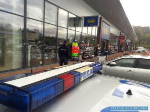 Policjant stoi na chodniku, na pierwszym planie dach policyjnego radiowozu