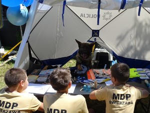 Policyjny namiot w którym znajduje się stół i ławki na których siedzi trójka chłopców oraz pies służbowy