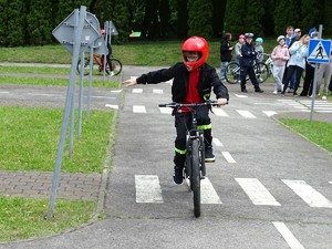 chłopiec jedzie na rowerze za nim stoi policjantka oraz grupa dzieci z rowerami