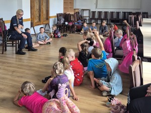 policjantka siedzi na krześle dookoła niej siedź, leżą  dzieci ma podłodze