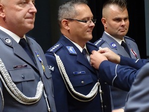 trzech policjantów w mundurach galowych, stojącemu w środku przypinany jest medal za zasługi