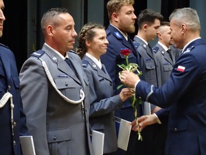 policjanci stoją szeregu w umundurowaniu galowym, policjantce policjant w mundurze stojący na przeciwko niej wręcza je ryżę i teczkę.