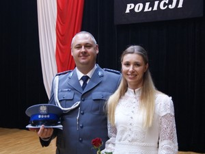 Policjant w mundurze galowym kobieta trzyma w ręku czapkę służbową i teczkę ,obok niego stoi w białej sukience trzyma w ręku różę