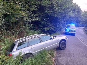 samochód marki audi znajduje się w przydrożnym rowie przód samochodu wystaje na pas jezdni na którym zaparkowany jest oznakowany radiowóz z włączonymi sygnałami świetlnymi
