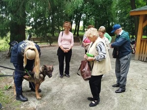 policjantka w mundurze zakłada szelki psu służbowemu w okół niej stoją seniorzy 5 kobiet i 1 mężczyzna