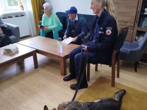 w pokoju na krześle siedzi policjantka w mundurze obok niej leży pies służbowy na kanapie obok siedzą seniorzy kobieta i mężczyana