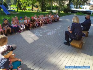 Umundurowani policjanci rozmawiają z grupa przedszkolaków. Wszyscy siedzą na ławkach na dworze, w tle plac zabaw. Policjanci siedzą na osobnej ławce po prawe stronie zdjęcia. Dzieci siedzą naprzeciwko funkcjonariuszy.