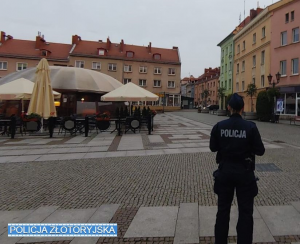 Na pierwszym planie stoi policjantka odwrócona plecami, w tle centrum miasta, kamienice mieszkalne i lokal gastronomiczny