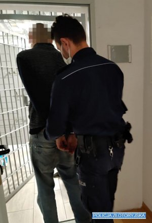 Policjant zakłada kajdanki na ręce zatrzymanego. Mężczyzna stoi przodem do krat, tyłem do policjanta. W tle areszt