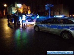 Nocą na ulicy stoi radiowóz policyjny, przed nim stoi samochód osobowy. Między pojazdami policjanci podejmują czynności wobec kilku osób.