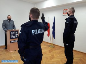 Policjanci w granatowych mundurach stoją tyłem, z podniesioną do góry prawą ręką. Przed nimi, za drewnianą mównicą stoi komendant w umundurowaniu wyjściowym. W tle flaga Polski i Unii Europejskiej oraz godło Polski