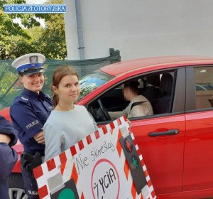 Policjantka z młodą dziewczyna stoją przy czerwonym samochodzie osobowym, w aucie za kierownicą siedzi kobieta, młoda dziewczyna trzyma w rękach plakat